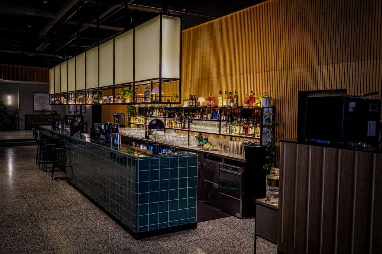 Modern bar in Fearns at Leeds Dock with a sleek green-tiled counter and a well-lit back wall displaying various bottles of liquor. The bar features a minimalist design with warm wooden panelling and overhead hanging lights, creating a cosy yet sophisticated ambiance.
