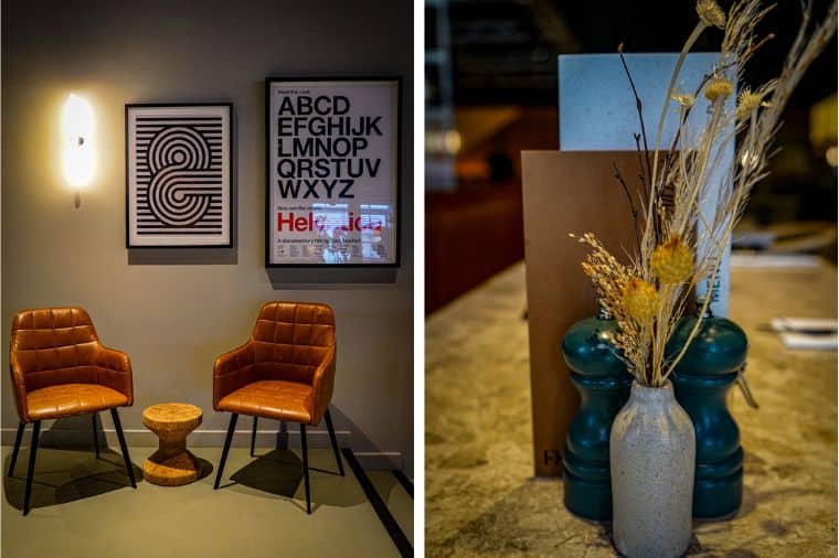 Split image featuring two scenes: on the left, two brown leather chairs with a small wooden side table between them, set against a grey wall adorned with framed typography art, including a poster of the Helvetica alphabet. On the right, a small vase with dried flowers and two green salt and pepper grinders on a table, next to a menu.