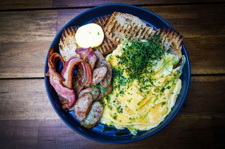 Folded eggs brunch dish from Fearns in Leeds Dock, featuring fluffy folded eggs topped with fresh herbs, served with crispy bacon, sliced sausage, and toasted sourdough bread with a pat of butter.