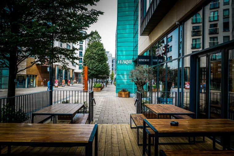 Outdoor patio seating area at Fearns in Leeds Dock, featuring wooden tables and benches overlooking a pedestrian walkway lined with trees. The Fearns sign is visible on the side of the modern building, with nearby planters adding greenery to the urban setting.